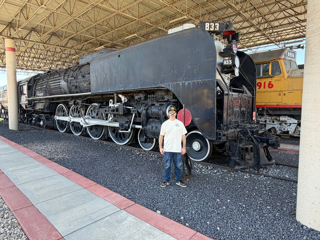 A man stands in front of a large black steam locomotive at a train museum under a covered platform, with another yellow train visible in the background.