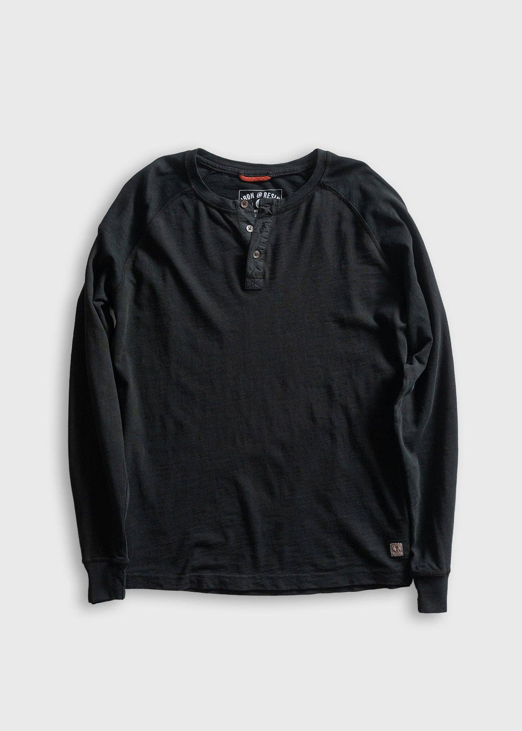 A black long-sleeve henley shirt with raglan sleeves, laid flat on a light gray background. The shirt features a three-button placket and a small brand patch near the bottom hem.