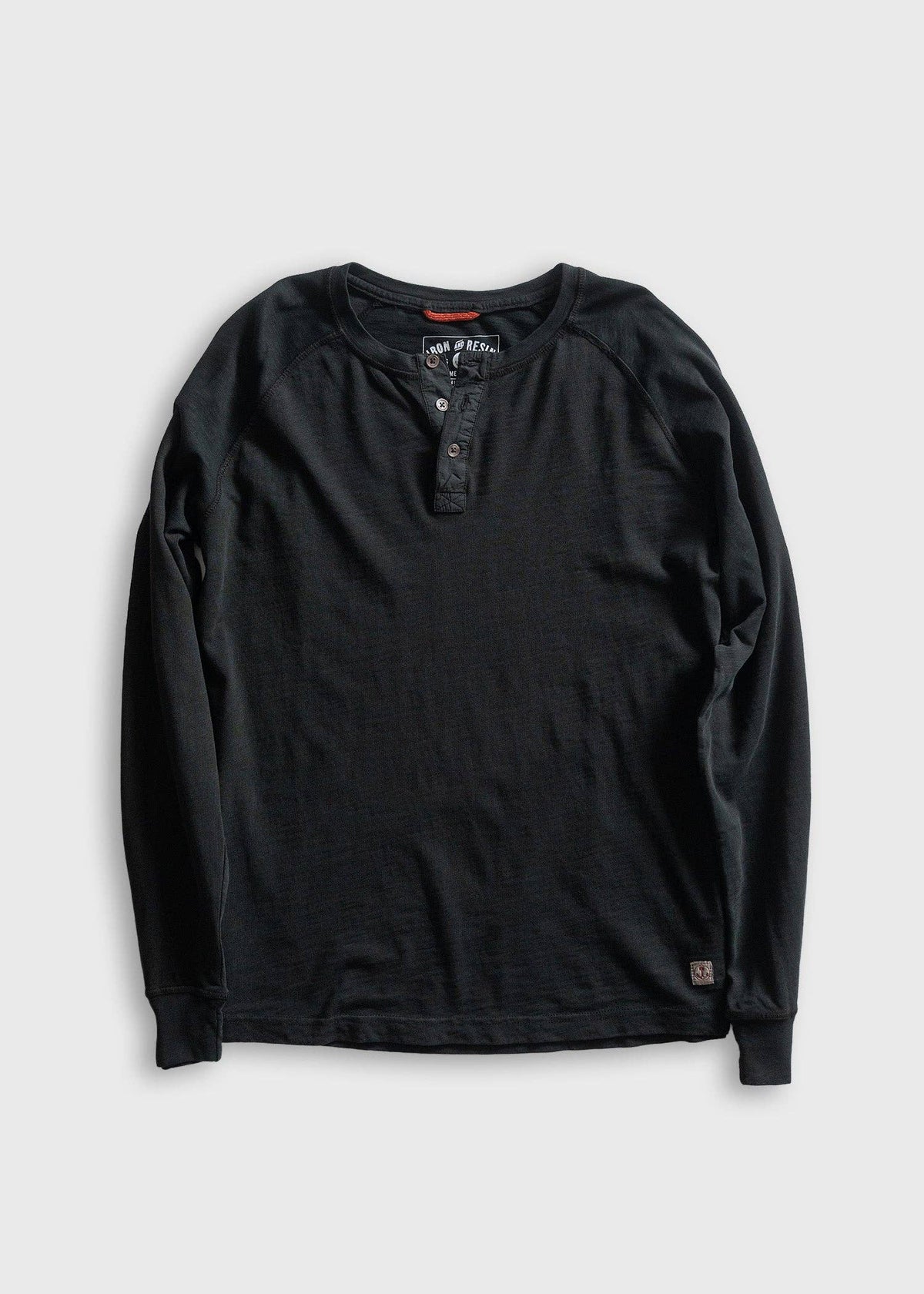 A black long-sleeve henley shirt with raglan sleeves, laid flat on a light gray background. The shirt features a three-button placket and a small brand patch near the bottom hem.