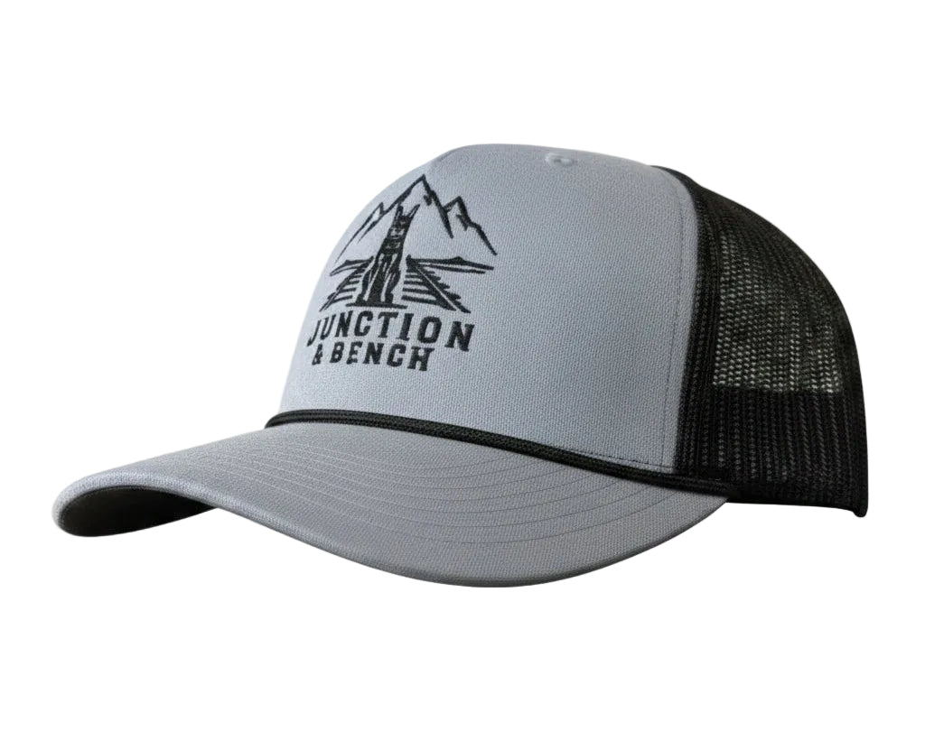 A light gray heather trucker hat with black mesh back panels and a black embroidered logo on the front that reads 'JUNCTION & BENCH' with a mountain and bridge graphic.