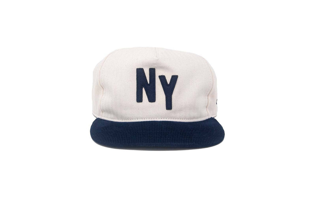 A structured 5-panel snapback cap with an off-white crown and a navy blue flat brim. The front of the crown features the letters 'NY' embroidered in navy blue.