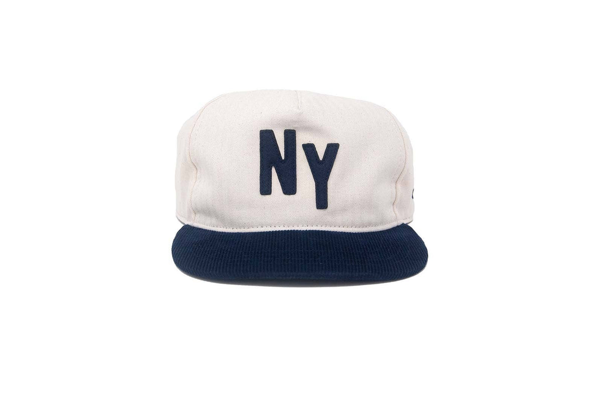 A structured 5-panel snapback cap with an off-white crown and a navy blue flat brim. The front of the crown features the letters 'NY' embroidered in navy blue.