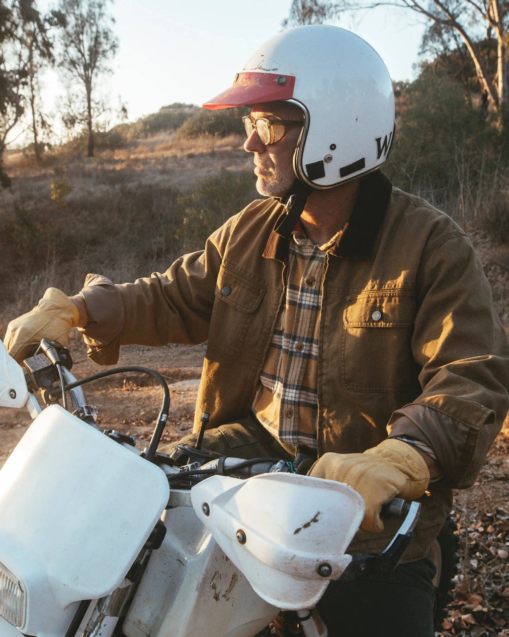 A man wearing a white vintage motorcycle helmet, sunglasses, a brown jacket, and tan gloves sits on a white dirt bike in an outdoor, dry grassy landscape during golden hour.