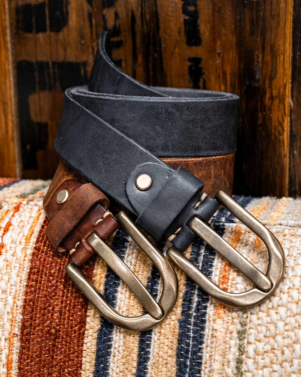Two leather belts, one black and one brown, with antique brass buckles, arranged on a striped woven textile in front of a rustic wooden background.
