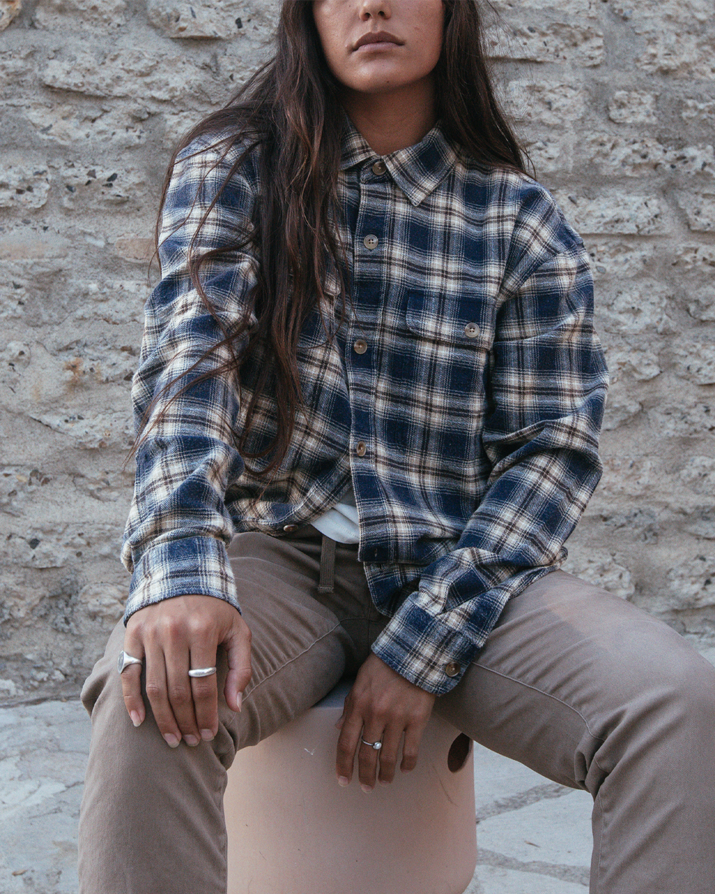 A woman wearing a navy, blue, and beige plaid button-up flannel shirt and taupe pants, sitting on a stool against a rustic stone wall.