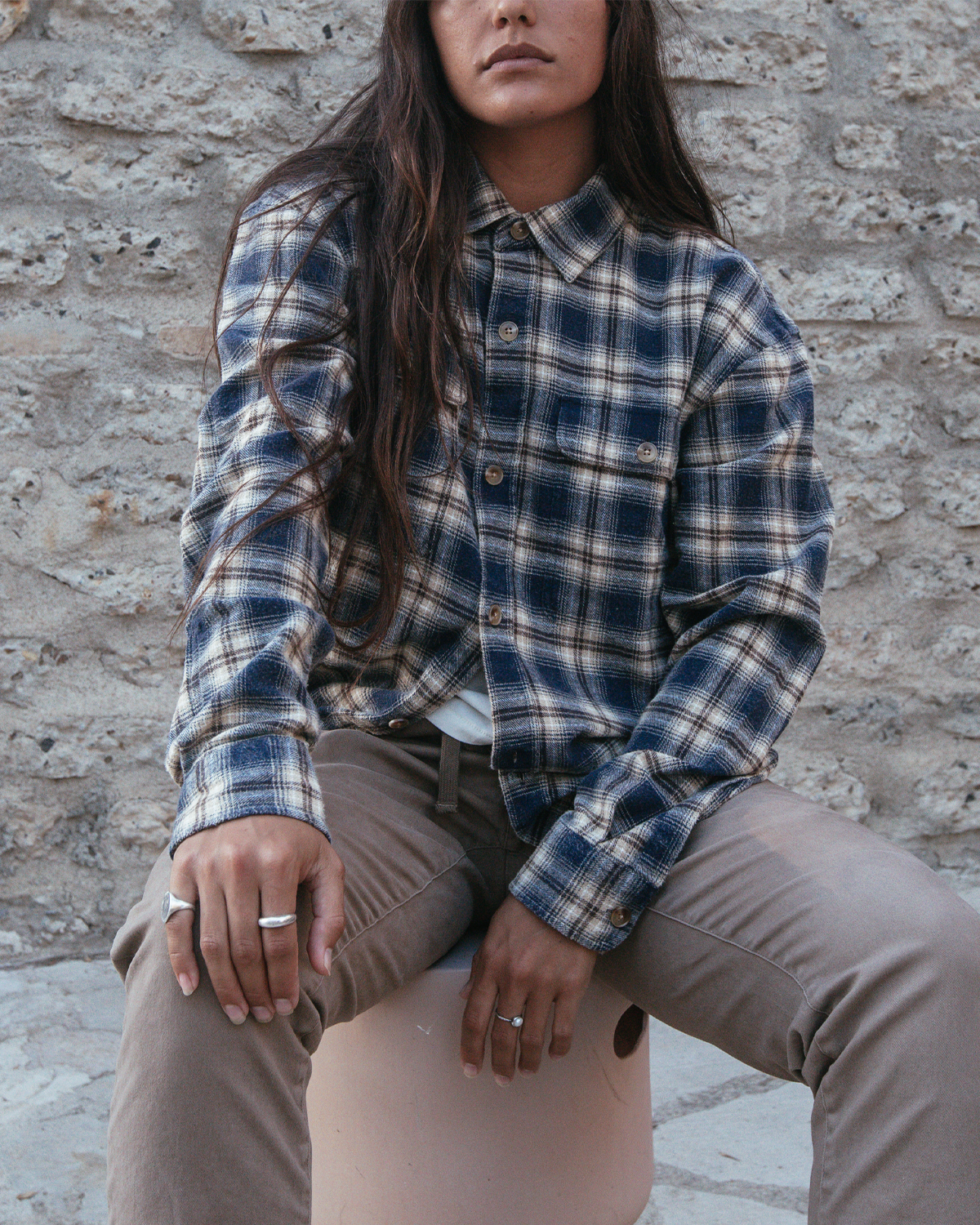A woman wearing a navy, blue, and beige plaid button-up flannel shirt and taupe pants, sitting on a stool against a rustic stone wall.