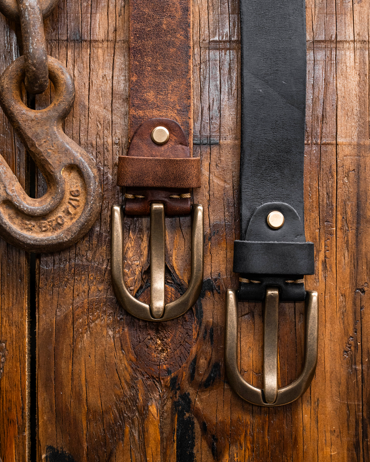 Two leather belts, one brown and one black, hanging on a rustic wooden plank background next to a rusty metal hook. Both belts feature antique brass double prong buckles and brass rivets.