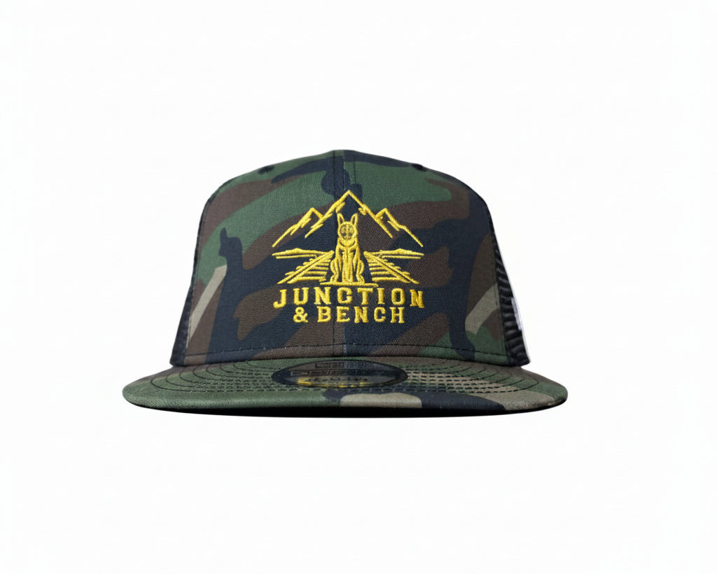 A camouflage trucker hat with a yellow mountain logo and 'JUNCTION & BENCH' text on the front, displayed on a wooden shelf against a textured beige wall.