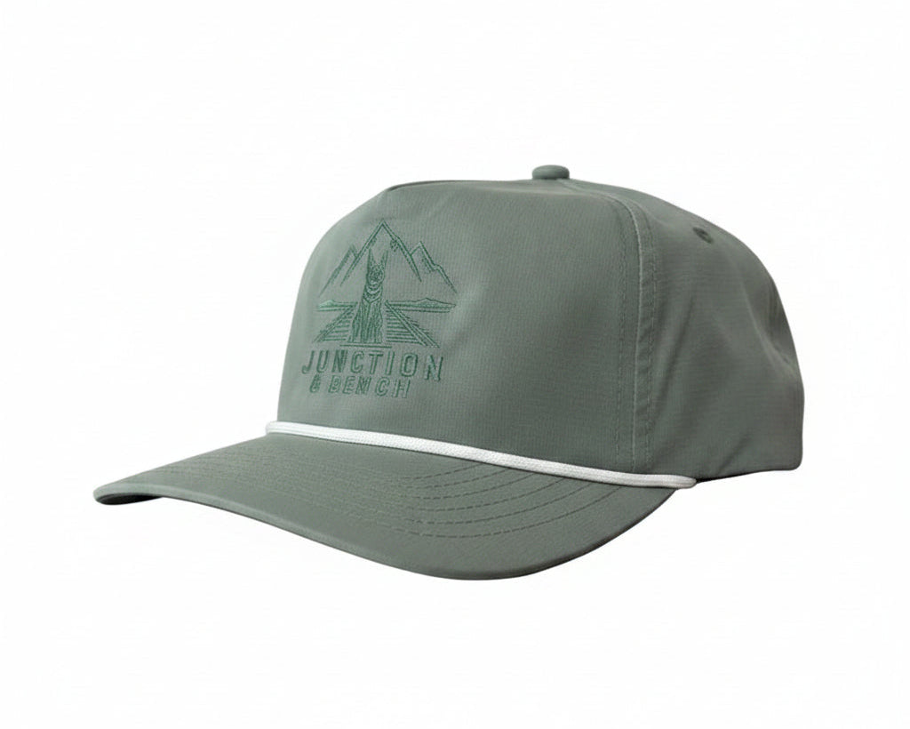 A sage green 5-panel trucker cap with a curved brim, featuring a tonal embroidered mountain and bridge logo with the text 'JUNCTION BENCH' on the front, and a thin white stripe across the brim.