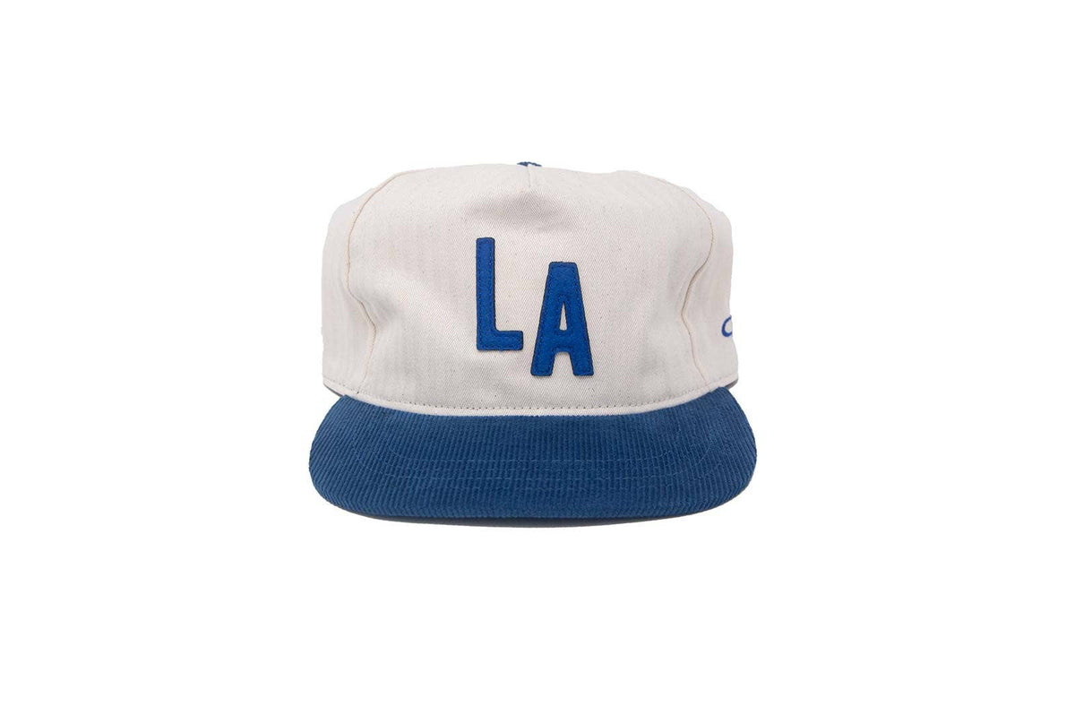 A white and blue LA snapback cap with a white crown, blue 'LA' lettering on the front, and a blue flat brim, shown against a plain white background.