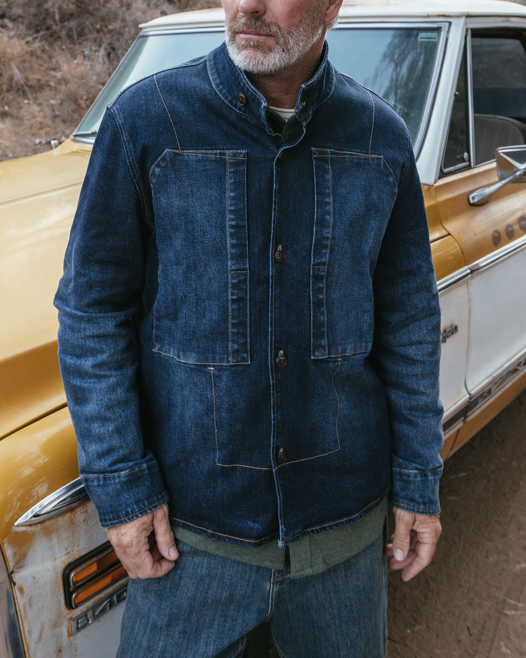 A man wearing a dark blue denim jacket with a stand collar, standing outdoors next to a vintage yellow and white truck.