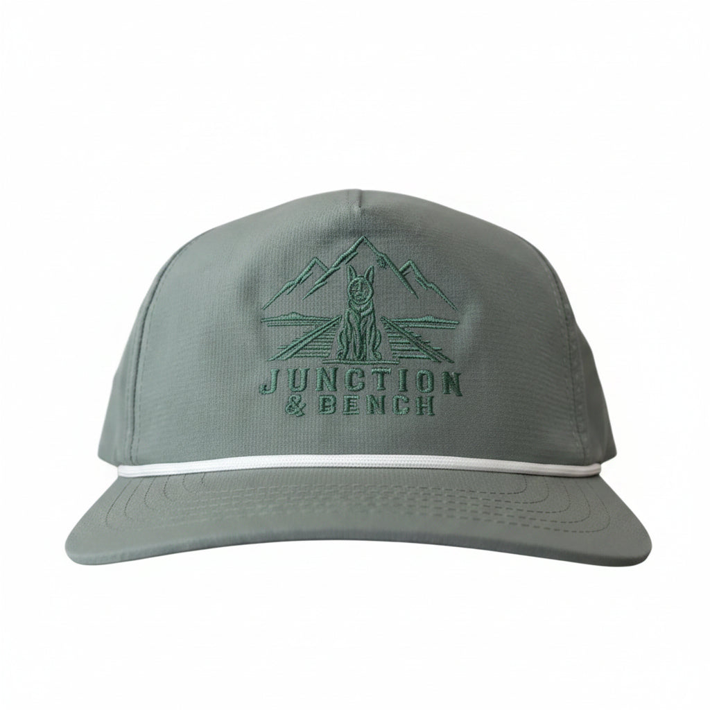 A light gray snapback cap with a green embroidered logo of mountains, a tree, and the text 'JUNCTION & BENCH' on the front, accented by a thin white stripe on the brim.