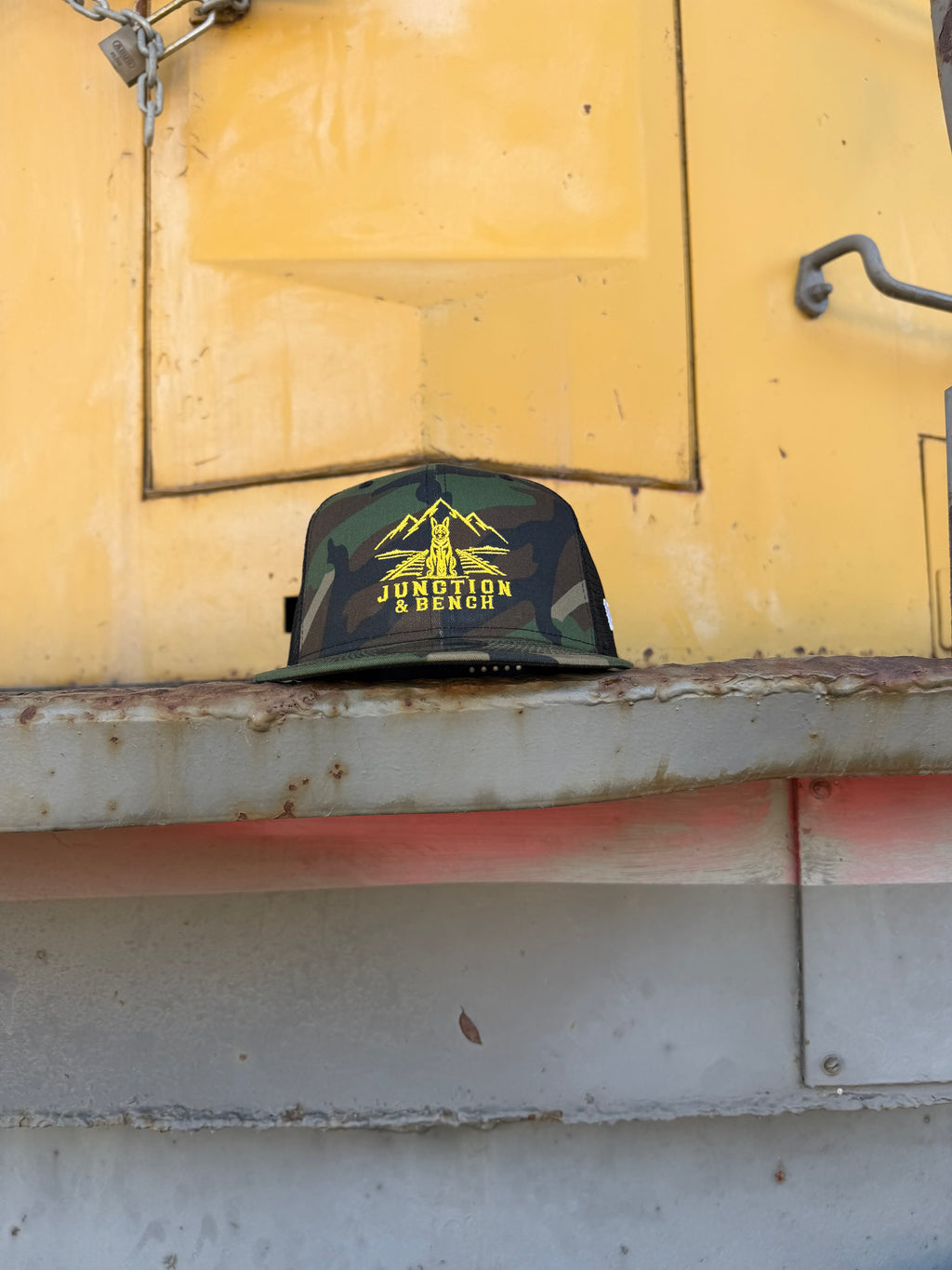 A camouflage baseball cap with a yellow 'JUNCTION BENCH' graphic and mountain logo, resting on a rusty metal ledge in front of a yellow industrial wall.