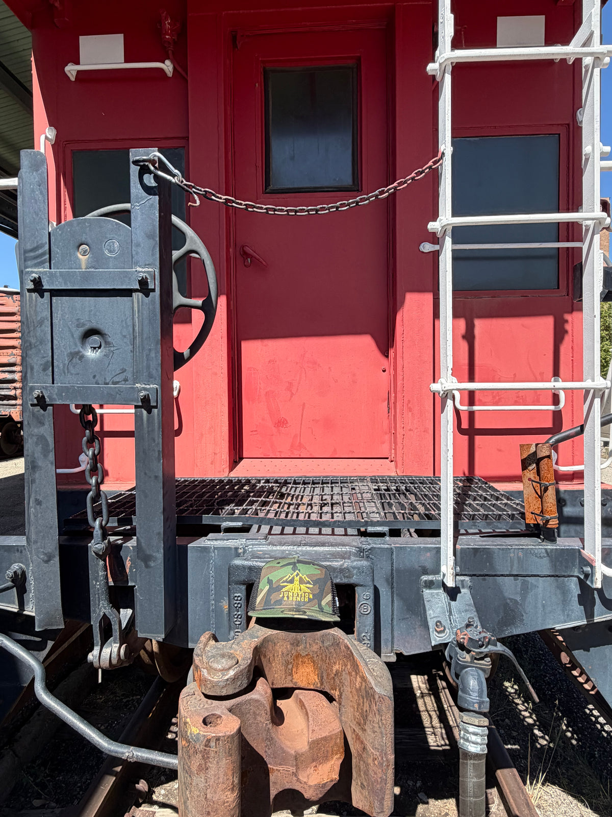 A close-up of a red train caboose with a black metal coupling device, rusty coupling hooks, a chain link across the doorway, and a white ladder on the right side.