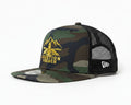 A camouflage trucker hat with a yellow 'JUNCTION BENCH' graphic on the front, displayed on a wooden shelf against a textured beige wall.
