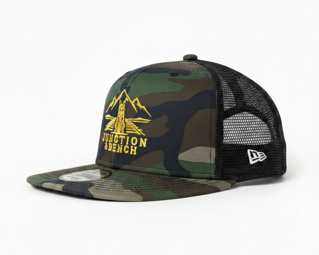 A camouflage trucker hat with a yellow 'JUNCTION BENCH' graphic on the front, displayed on a wooden shelf against a textured beige wall.