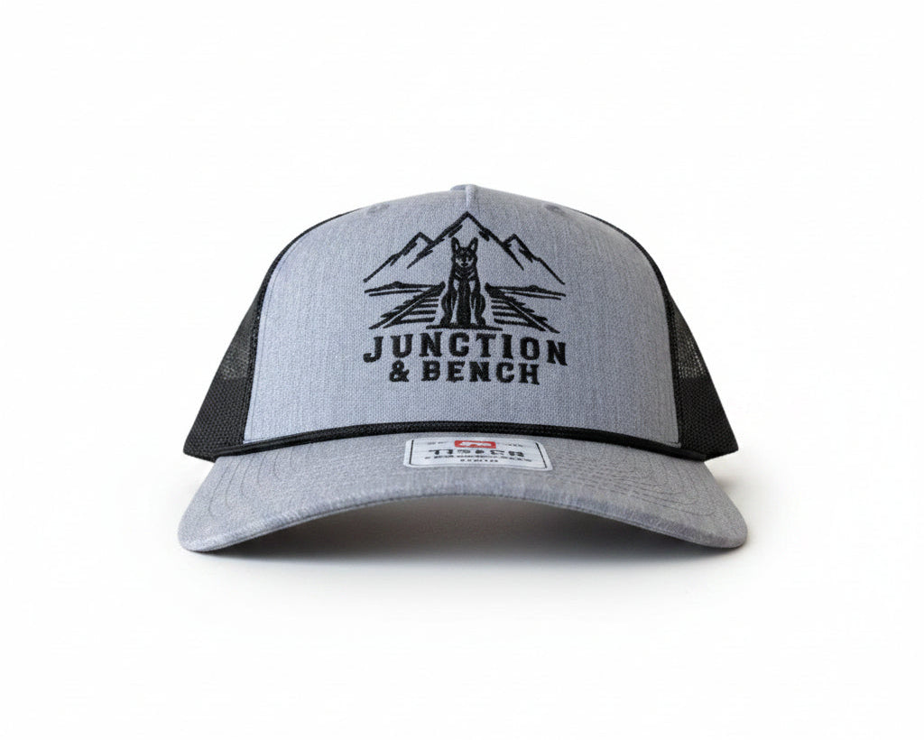 A light gray and black trucker hat with a black embroidered logo of mountains, a wolf, and the text 'JUNCTION & BENCH' on the front panel. The hat has a heather gray front and bill, with black mesh back panels.