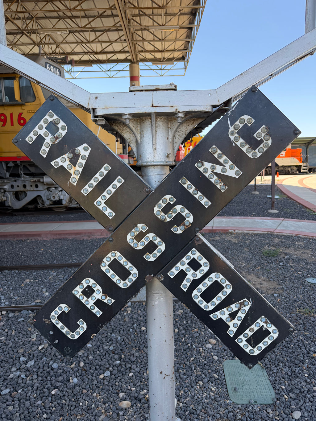 A railroad crossing signpost with four black rectangular signs spelling 'RAILROAD CROSSING' in white letters, each outlined with small light bulbs. The sign is mounted on a metal post at a train station, with gravel tracks, a train, and a station canopy visible in the background.