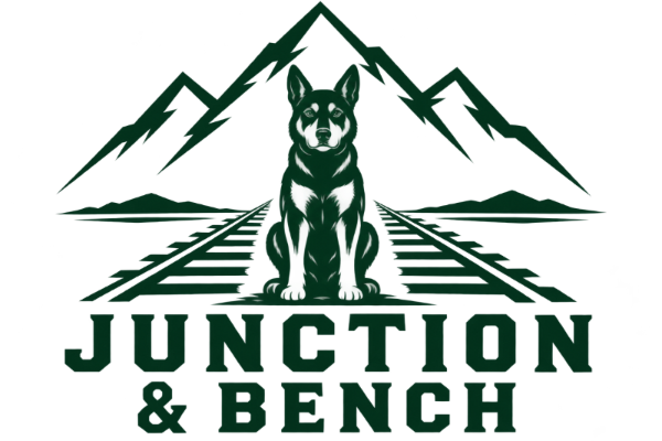 A dark green and white graphic logo featuring a seated dog centered on railroad tracks, with jagged mountains above and the text 'JUNCTION & BENCH' below.