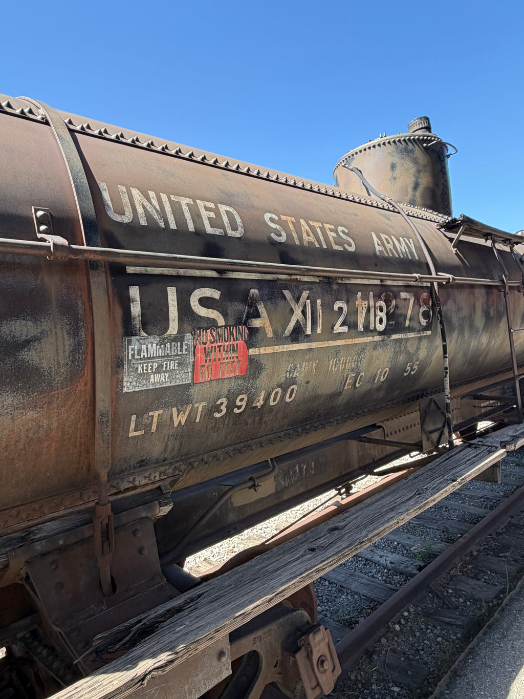 Close-up of a weathered brown and black United States Army tank car on railroad tracks under a clear blue sky, with white lettering and a small red warning label.