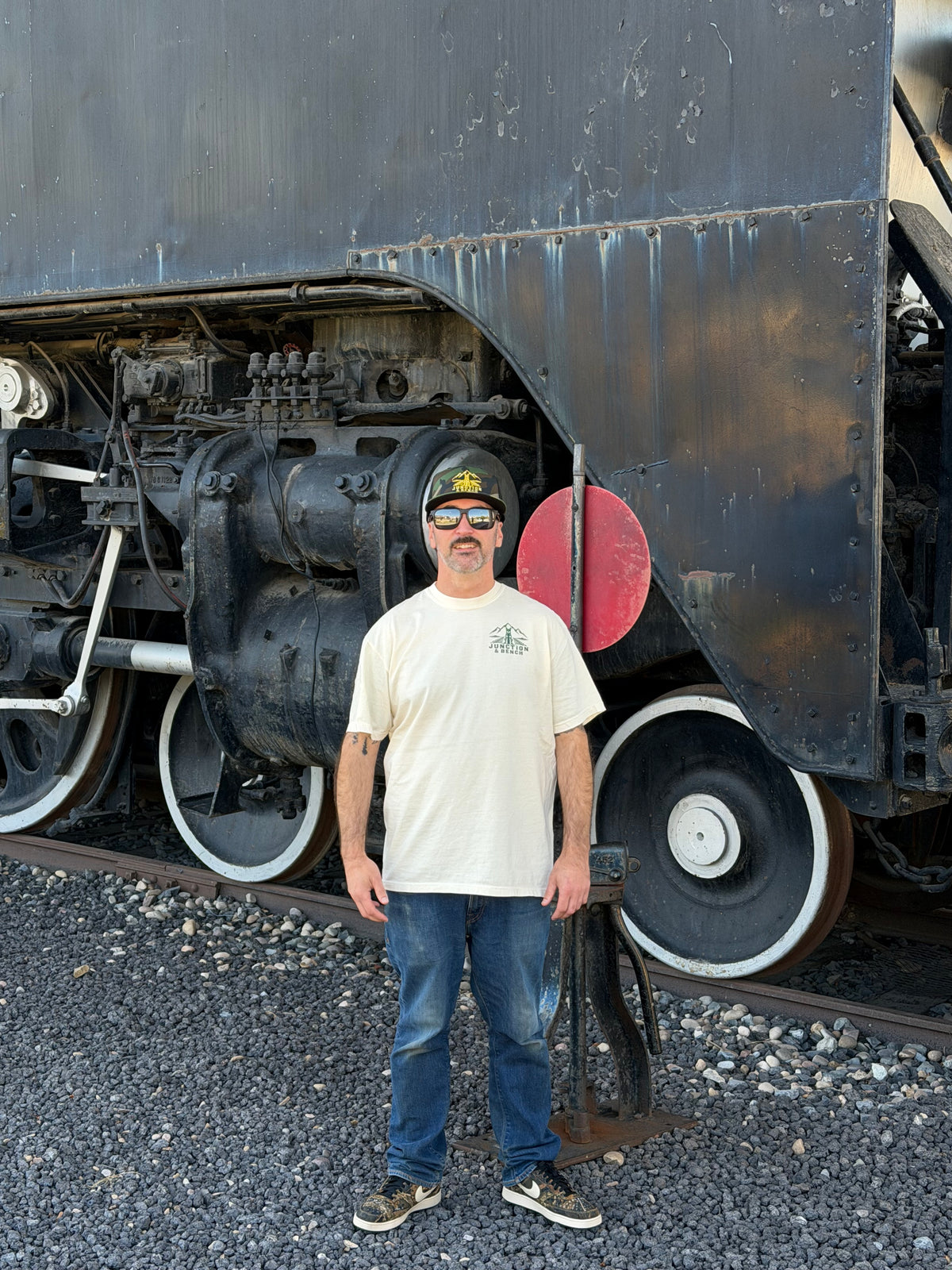 A man wearing a cream t-shirt, blue jeans, and a black cap stands in front of a large vintage black steam locomotive on a gravel track.