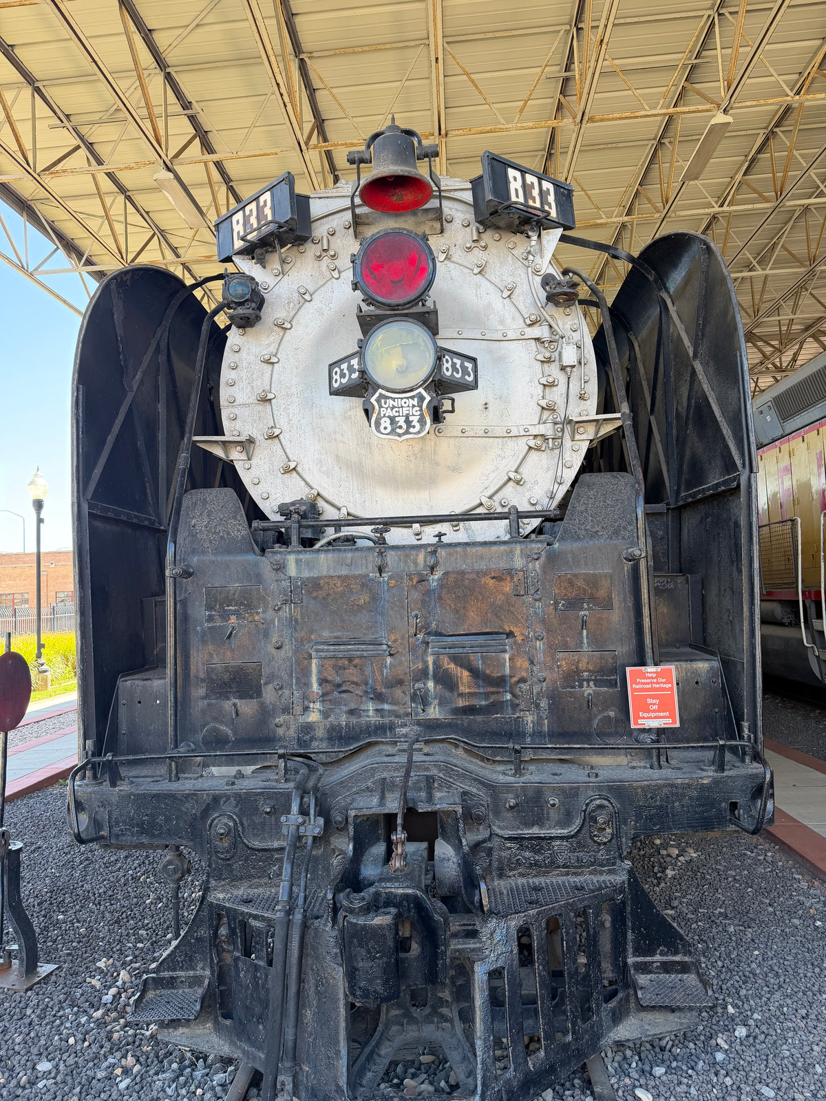 Front view of a vintage steam locomotive train under a covered platform, with a black boiler and tender, white front cab, and red and white lights.