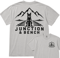 A light gray short-sleeve t-shirt shown from the back, featuring a large black graphic of a dog sitting on railroad tracks with mountains behind it and the text 'JUNCTION & BENCH'. A smaller inset at the bottom right shows the front chest print of the same design.