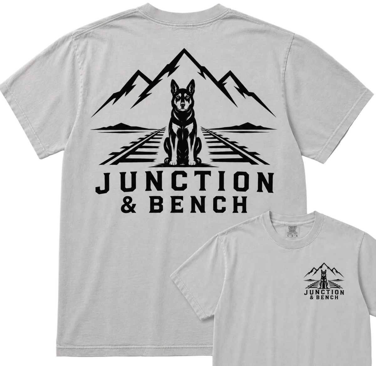 A light gray short-sleeve t-shirt shown from the back, featuring a large black graphic of a dog sitting on railroad tracks with mountains behind it and the text 'JUNCTION & BENCH'. A smaller inset at the bottom right shows the front chest print of the same design.