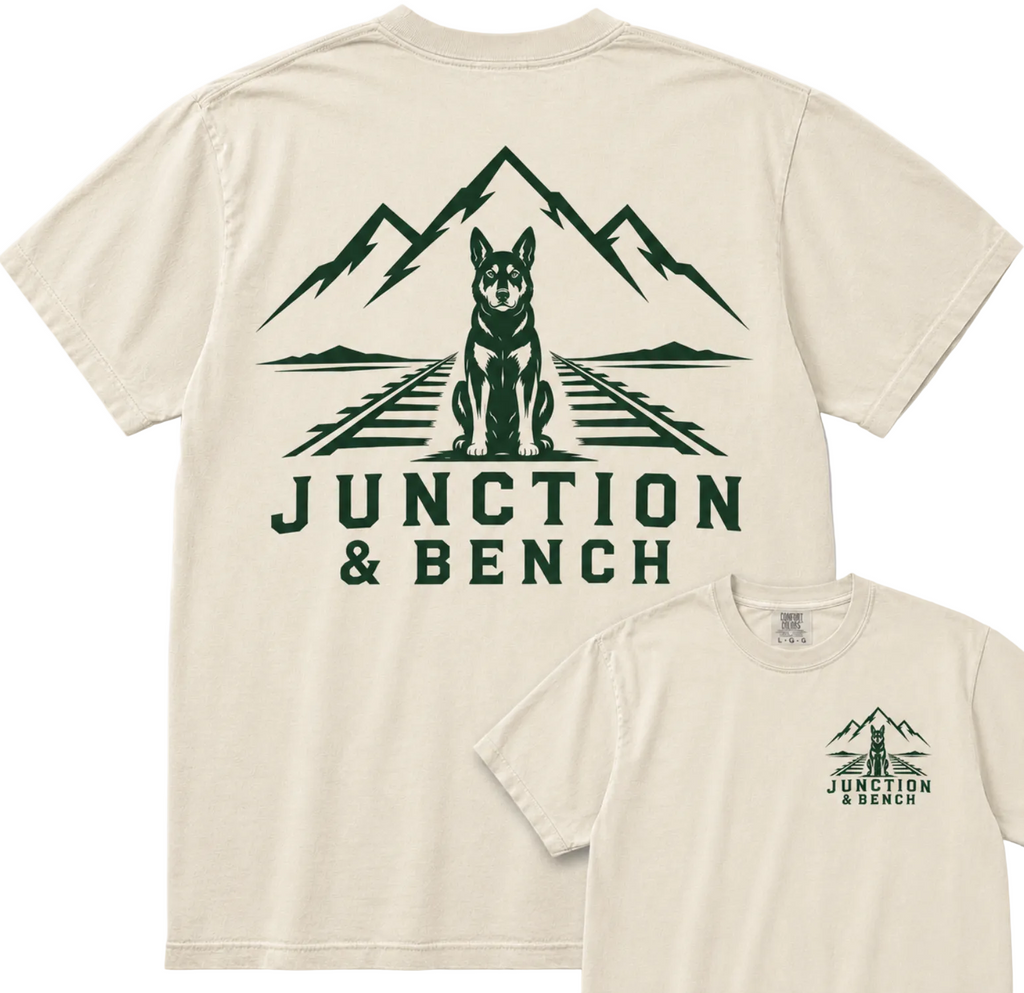 Two cream-colored t-shirts showing the front and back designs. The back shirt features a large dark green graphic of a dog sitting on train tracks with mountains in the background and the text 'JUNCTION & BENCH'.