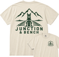 Two cream-colored t-shirts showing the front and back designs. The back shirt features a large dark green graphic of a dog sitting on train tracks with mountains in the background and the text 'JUNCTION & BENCH'.