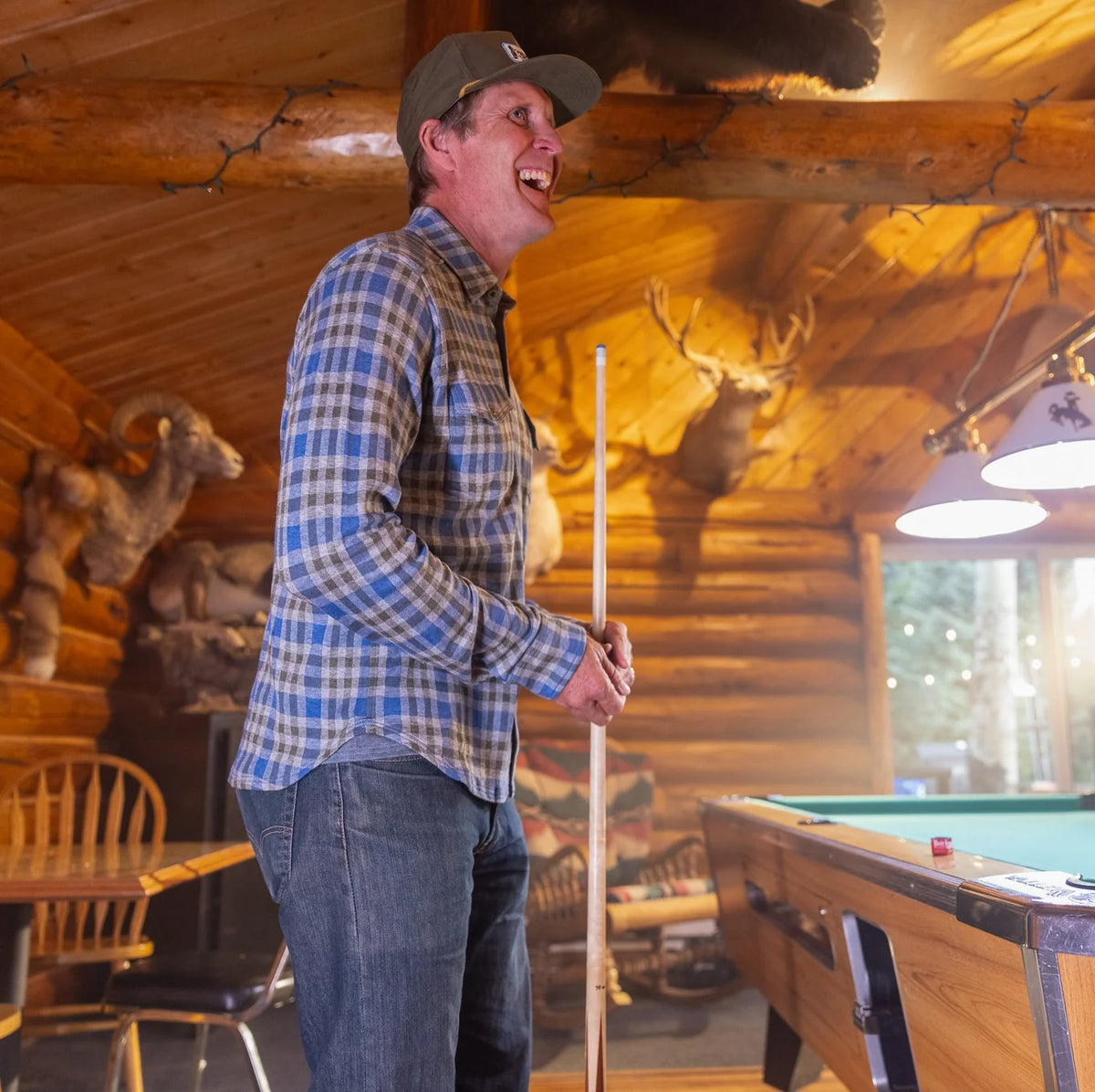 A man in a blue and gray plaid flannel shirt and jeans stands in a rustic log cabin holding a pool cue, smiling in a room with a pool table, mounted deer heads, and warm wood lighting.