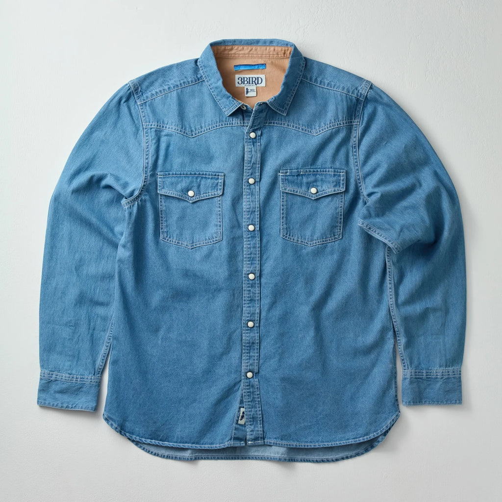 A blue denim long-sleeve button-up shirt with two chest pockets, laid flat on a light gray background.