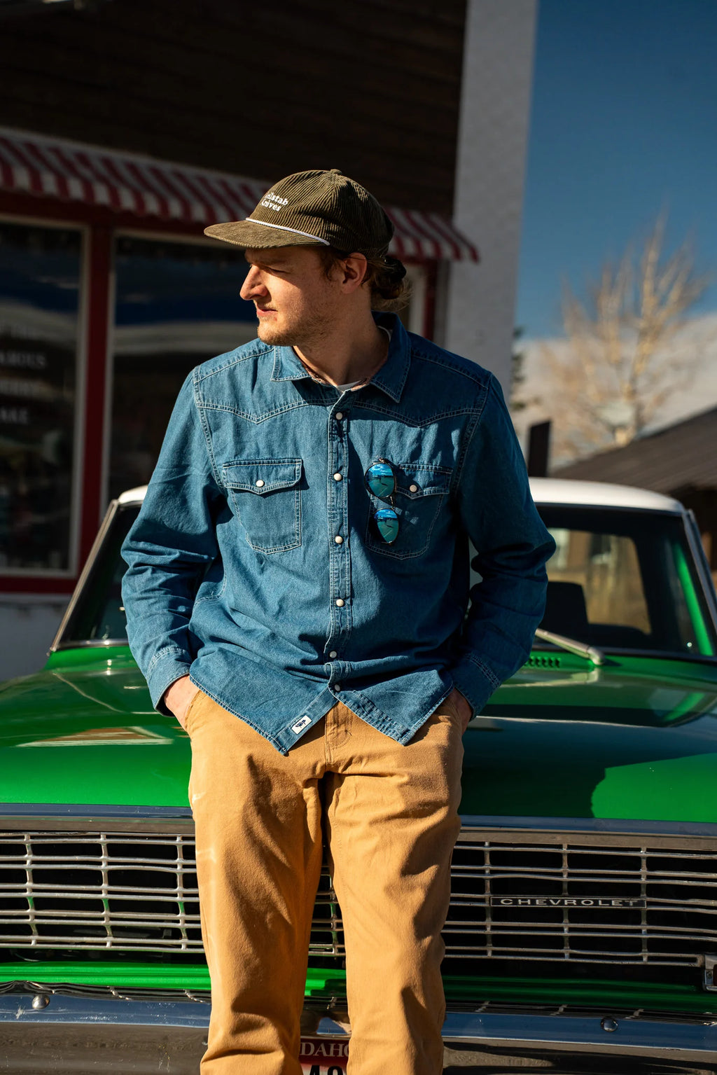A man wearing a blue denim button-up shirt, tan pants, and a green cap stands in front of a vintage green Chevrolet truck outdoors.