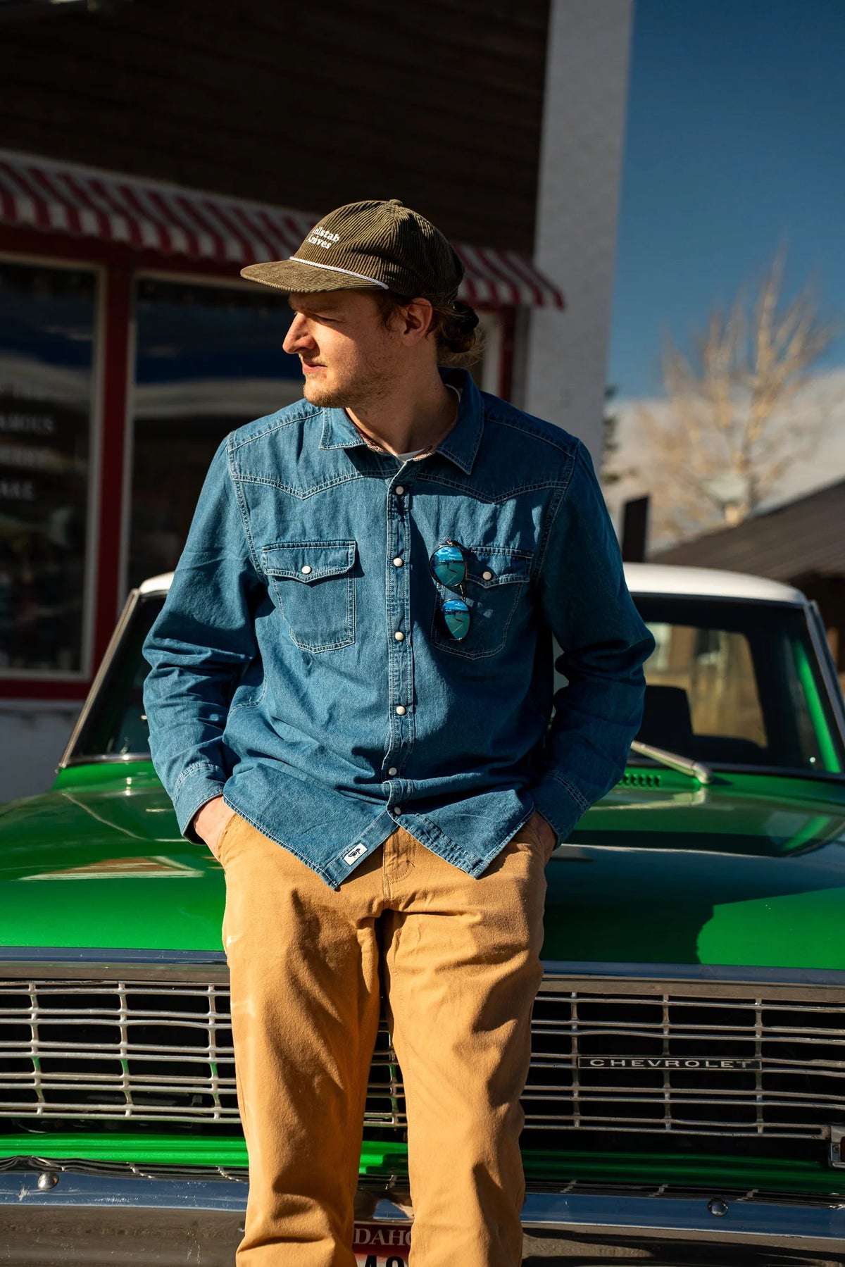 A man wearing a blue denim button-up shirt, tan pants, and a green cap stands in front of a vintage green Chevrolet truck outdoors.