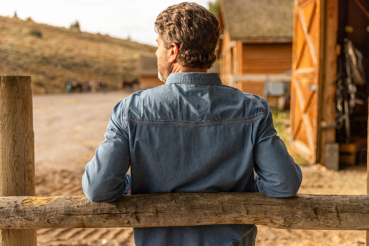 Man in a blue shirt leaning against a wooden fence in a rural setting with a barn and horses in the background.