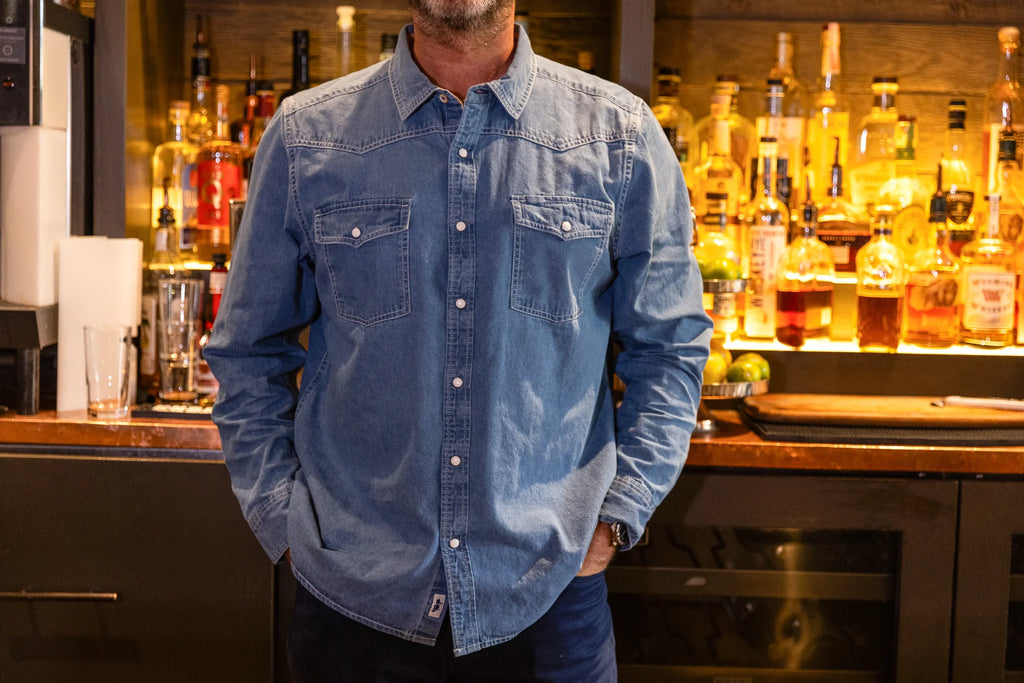 Person wearing a blue denim shirt standing in front of a bar with bottles in the background