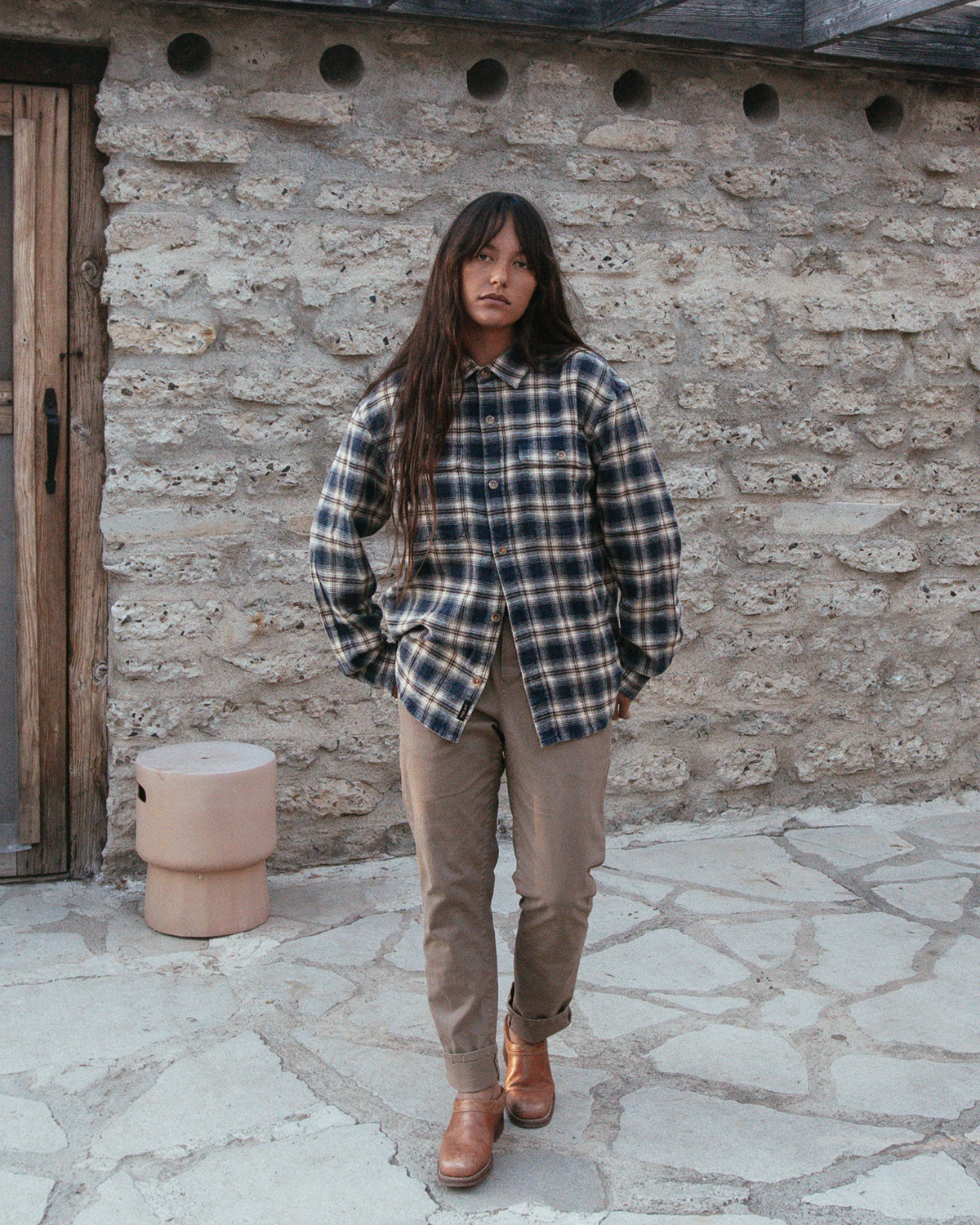 A woman stands outdoors in front of a rustic stone wall, wearing a dark blue and brown plaid flannel overshirt, tan pants, and brown leather boots.