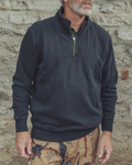 A man wearing a dark charcoal gray quarter-zip pullover sweater with a mock neck and zippered chest pocket, standing against a rustic stone wall.