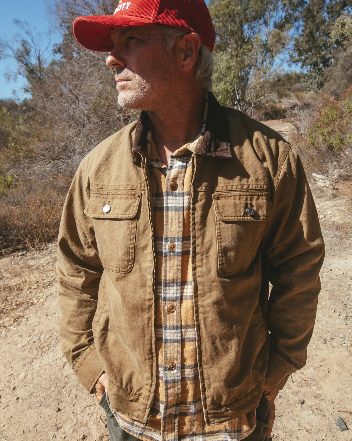 A man wearing a brown corduroy utility jacket over a tan and blue plaid flannel shirt, with a red baseball cap, standing outdoors on a dirt path with trees in the background.